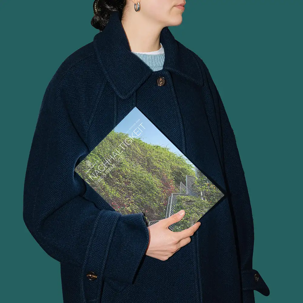 A person wearing a navy blue coat holds a magazine with a cover image showing lush green vines growing on a staircase. The scene conveys the theme of sustainability.